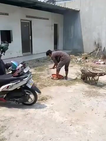 A person wearing a brown shirt and pants is breaking up cement with a shovel in a dirt and gravel area. The person is standing over a circular concrete mold and is using the shovel to chip away at the cement. A motorcycle is parked to the left of the frame, and a wheelbarrow filled with cement is to the right. A single-story building with two doors is visible in the background.