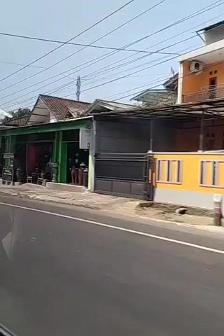 The video shows a street scene with shops lining the right side of the road. The shops are painted in various colors, including green, white, and grey. There are signs in front of some of the shops, one of which reads "BAKSO MIE AYAM." People are seen riding motorcycles and walking on the sidewalk. The weather appears to be sunny with a clear sky. Overhead, power lines are visible. The road is paved and appears to be a two-lane road. There are some potted plants and trees in front of the shops.