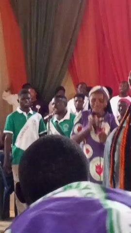 The video shows an indoor gathering with a stage decorated with draped fabric in shades of green and red. A woman in a purple dress with red heart designs is speaking into a microphone. She is wearing a headscarf. Several other people are visible in the background, some wearing green and white uniforms. The audience is partially visible in the foreground, with their backs to the camera, some wearing purple and white attire. The overall impression is of a formal event or ceremony.