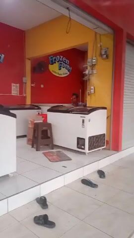 The video shows the interior of a frozen food shop. The walls are painted red and yellow. There are several white chest freezers in the shop. A person is visible behind a counter in the back of the shop. There are two signs that say "Frozen Food". A brown stool is placed in front of one of the freezers. Several pairs of black flip-flops are scattered on the tiled floor outside the shop entrance.