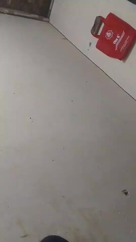 The video begins by showing a corner of a room. The wall is painted a light color and appears to be slightly dirty. In the upper right corner of the frame, a red tote bag with a white logo is hanging on the wall. Above the tote bag, some clothing, including a blue and white striped shirt and a black shirt, is visible. The floor is also light-colored and shows some signs of wear and tear, with a few dark spots and a small hole. The lighting in the room is dim, casting shadows and making it difficult to see clearly. The camera angle is tilted downwards, giving a somewhat distorted view of the space.