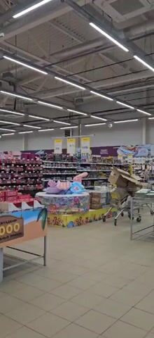 The video pans across a well-lit grocery store aisle, showcasing a variety of candy bars. The camera begins on the left, showing rows of boxed chocolates such as Snickers and M&Ms. It then moves to the right, revealing more candy displays, including Twix and Milky Way bars. In the center, there is a display featuring inflatable toys. The aisle is lined with shelves stocked with various products, and a shopping cart is visible on the right side of the frame. A promotional sign with Russian text and an image of a beach can be seen in the foreground. The lighting is consistent throughout the scene, indicating an indoor environment.