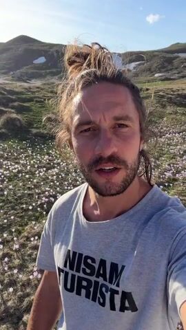 A man with shoulder-length brown hair in a bun and a beard is holding a phone and filming himself. He is wearing a gray t-shirt that says "NISAM TURISTA." He is standing in a field of tiny white and light purple flowers. Behind him is a gently sloping field with some brown vegetation and a small pond in the distance. The sky is mostly blue.  He looks at the camera and talks. Then he turns the camera up towards the sky, and falls backwards into the flowers. He looks up at the sky. Then he rolls over and stands up again, facing the camera. He points at the camera. He makes an “ooh” sound.