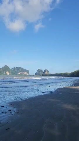 The video shows a beach with dark sand and shallow water. The water has ripples and reflects the sky. There are some pieces of driftwood on the sand. In the background, there are large, green, mountainous islands in the distance. The sky is blue with some white clouds. The waves are gentle and break on the shore. The scene appears to be a natural landscape with no visible people or man-made structures other than what could be natural debris. The overall impression is of a serene and natural coastal environment.