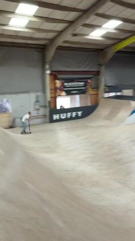 Here's a description of the video:
The video shows a person riding a scooter in an indoor skate park. The person, wearing a white t-shirt and jeans, begins on a flat, wooden surface before riding up a wooden ramp, gaining height and momentum. They then transition to a quarter-pipe style ramp with a black surface, riding up the curved surface before presumably turning around out of view. The skate park environment includes various ramps, wooden surfaces, support beams, and banners on the walls.