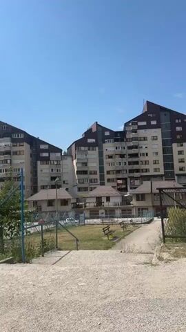The video shows a series of apartment buildings with distinctive angular roofs against a clear blue sky. In the foreground, a chain-link fence runs horizontally across the frame. Beyond the fence, there are patches of grass, a small tree, and a wooden bench. Further back, a few cars are visible, parked or driving in what appears to be a parking lot or street area. The buildings are multi-story, with numerous windows and balconies. The overall environment appears to be a residential urban area on a sunny day.