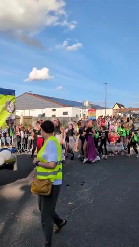 This is a vertical video of a group of young women in colorful skirts and black tops dancing in a procession. They are walking down a street, and the camera is following them from behind. The women are smiling and appear to be enjoying themselves. The background shows buildings, trees, and a blue sky with clouds. There are other people watching the procession from the side of the street. The video is well-lit and the colors are vibrant. The overall tone of the video is festive and upbeat.