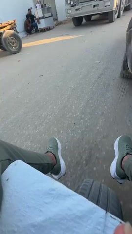 A video shows a person's legs and feet, wearing green pants and white sneakers, sitting on what appears to be the edge of a tire or a vehicle. The camera pans across a road with cracked asphalt. To the left, two people are visible in the background; one is sitting on the ground, and another is standing. Further ahead, a large red truck is parked, with its front visible. The truck has the Volvo F12 logo on it and appears to be carrying a large, tarp-covered load. The video continues to pan to the right, revealing more of the truck's side and its multiple wheels. The lighting suggests it is daytime, with a clear sky. The overall scene appears to be an outdoor setting, possibly a roadside or a staging area.