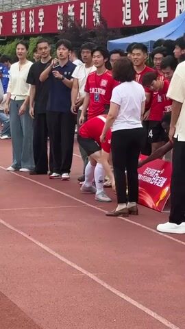 Here's a detailed description of the video:

The scene takes place on a red running track, likely at a sports field associated with a university in China. A group of young men, mostly wearing red athletic shirts and shorts, are gathered together. Some of them are huddling in a circle with their arms around each other's shoulders. One man is bent over and appears to be stretching or receiving assistance from a woman dressed in a white top and black pants. She is standing beside him, seemingly guiding or supporting him. Another man is holding a microphone and appears to be speaking or announcing something, while others look on. Several other individuals, including spectators and staff, are standing around the edges of the group, observing. 

In the background, there's a large banner in Chinese.  Also visible are buildings, greenery, and a few people sitting in the upper levels of a structure that seems to be part of the sports complex. There is also a small blue tent behind the crowd. The weather appears to be overcast.