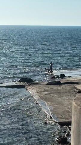 The video shows a view from a concrete structure looking out towards the ocean. Two people are standing in the shallow water near the structure's edge. One person is wearing a hat and dark clothing, while the other is wearing what appears to be swimwear. The ocean is blue with small waves, and the sky is clear and bright. The video seems to have been taken during a sunny day.