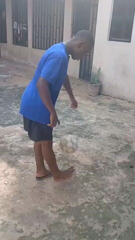 The video shows a young man with dark skin and short black hair playing with a soccer ball in a courtyard. He is wearing a bright blue t-shirt with a logo on the back and dark shorts. He is barefoot, except for one flip-flop on his right foot. He skillfully kicks the ball up into the air with his right foot, then with his left, and continues to keep it airborne with his feet and legs.

The scene appears to be a residential area in Egbagu, Nigeria. The ground is a rough, concrete surface. In the background, there is a building with barred windows and a large, wooden structure leaning against it. A potted plant sits near the building. The lighting suggests it is daytime, likely morning or afternoon. The man's focus and controlled movements indicate he is enjoying the activity. There is no visible text in the video.
