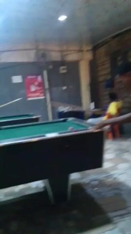 A man in a blue baseball cap, denim shirt, and dark trousers stands behind a green pool table, holding a pool cue. The table has several billiard balls on it. Another person is visible in the background to the left, partially obscured, standing at another pool table. The room has dark gray walls with advertisements posted, a drop ceiling with fluorescent lighting, and a tiled floor.  The camera is positioned low, looking up towards the pool table and the man.