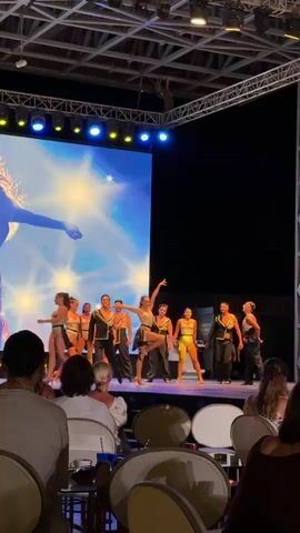 This media file captures a performance taking place on a stage under a night sky. The stage is lit by a large screen displaying a silhouette of a woman with flowing hair against a bright, star-like background. In front of the screen, a group of dancers in shimmering outfits, a mix of leotards and pantsuits, are performing. They appear to be a mix of male and female performers. The dancers are dressed in gold, silver, and black costumes. Some dancers are striking poses, while others are in motion.

The scene is viewed from the perspective of an audience seated in chairs in what appears to be an outdoor venue, possibly a resort or an amphitheater. The audience members are mostly visible from behind, showing their backs and heads. The lighting suggests it is nighttime, with stage lights illuminating the performers and the backdrop. The overall atmosphere seems lively and celebratory, with the performers exuding energy and confidence. There is no visible text in the frame.