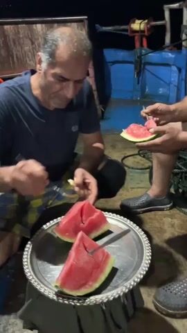 Three men are on a boat at night, eating watermelon. One man in a navy blue t-shirt and plaid shorts is sitting down and using a fork to eat a slice of watermelon from a metal platter. Another man, shirtless, is sitting and holding a slice of watermelon. A third man, wearing a blue tank top and plaid shorts, is also sitting and holding a slice of watermelon. The boat appears to be at sea, with a dark sky and water visible in the background. There are various ropes and equipment on the boat. A plate of flatbread is visible in the foreground.