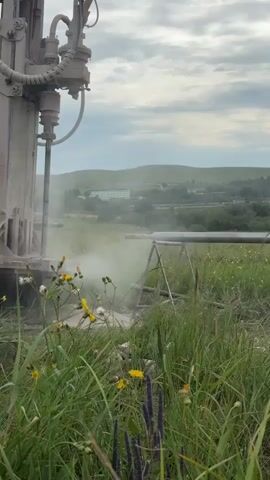A heavy-duty drilling rig is in operation, its drill bit actively penetrating the ground. Dust and debris are being ejected from the borehole in a plume of white. The rig is situated in a grassy field with wildflowers scattered throughout. In the background, rolling hills are visible, dotted with buildings and trees, suggesting a rural or semi-urban environment. The sky is overcast with scattered clouds. The overall scene depicts an industrial or construction activity taking place outdoors in a natural setting.