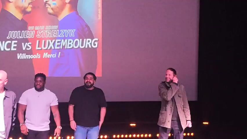 The video shows four men standing on a stage in front of a large screen. The screen displays an image of two men and text that reads "On Man Show, Julien Strelzyk, France vs Luxembourg, Vilmools Merci!". The men on the stage are smiling and appear to be interacting with each other. The man on the right is wearing a jacket and adjusting his collar. The stage is dark with several small lights arranged along the front edge.