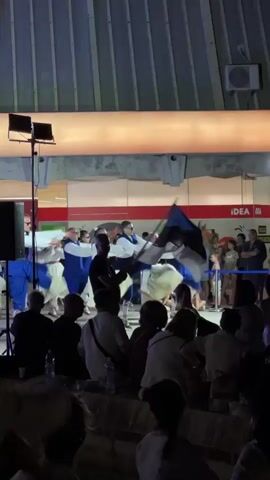 A group of dancers in white dresses and blue vests perform a coordinated dance. One dancer in the center holds up a flag with blue and white stripes. The dancers are on a stage in front of a building with a sign that says "IDEA". An audience is seated in the foreground watching the performance.