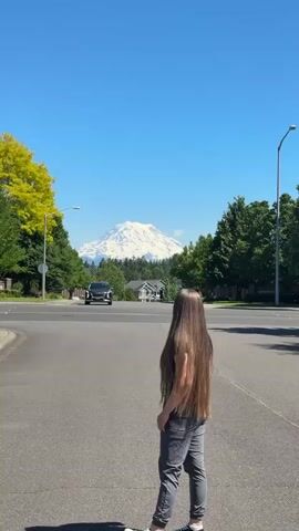 A young woman with long brown hair runs towards the camera on a paved street. She is wearing a black t-shirt and gray pants. Behind her, a snow-capped mountain is visible in the distance, with trees and houses on either side of the street. A car is driving down the street in the background. The sky is clear and blue.