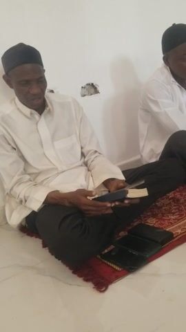 Two men are sitting on the floor against a white wall, on a red patterned rug. The man in the foreground is holding a book, with his hands resting on his lap. He is wearing a white shirt, dark pants, and a small dark cap. The man in the background is wearing similar attire, but the top of his head is out of view. Several small black objects are placed on the rug near the first man. A small damaged area is visible on the white wall.