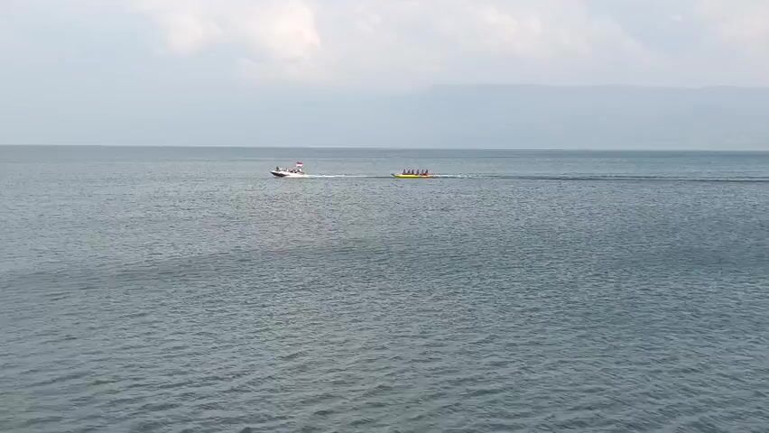 A motorized boat tows a banana boat with several passengers across a large body of water under an overcast sky. The boat is white with red trim and a flag at the stern. The banana boat is yellow and is shaped like a banana. The water is choppy, and there is land visible in the distance on the left side of the frame. The scene appears to be a recreational activity on a lake or sea.
