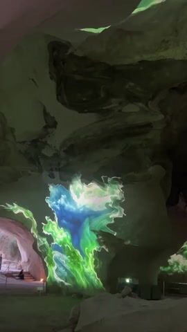The video shows a large cavern with projections of vibrant, abstract colors and patterns on the walls and ceiling. The projections shift and flow, creating a dynamic visual experience. In the foreground, large rocks are visible. In the background, there are some stairs and a railing, suggesting a pathway for visitors. A person can be seen sitting on the stairs in the lower left portion of the frame. The cavern walls are rough and textured, with some areas appearing smooth and others more jagged. The lighting is dim, except for the projected colors, which illuminate the space.