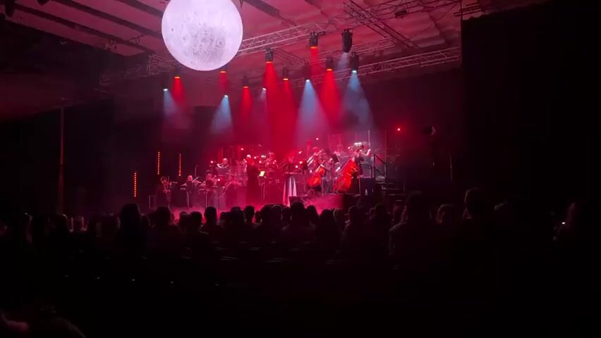A concert hall is lit in red, with a full orchestra playing on a stage. A conductor stands in front, leading the musicians. A large, white, spherical moon prop hangs above the stage, with spotlights focused on the orchestra. The audience sits in the dark, watching the performance.