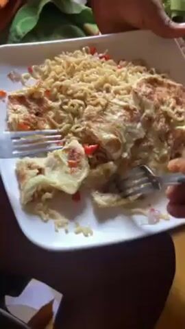 The video shows a person or persons using two forks to divide a dish of noodles and what appears to be an omelet or fried egg on a white square plate. The noodles are light brown and interspersed with diced red and white vegetables. The omelet has a slightly browned surface and contains similar diced vegetables. The person holding the plate has dark skin. In the background, there is a green patterned fabric and a glimpse of a wooden surface.