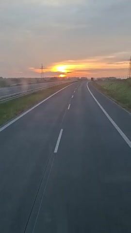 This video shows an empty highway during sunrise. The sun is low on the horizon, casting a warm orange glow across the sky. The highway is paved and has white lines marking the lanes. There are power lines and vegetation visible in the background. The camera appears to be moving forward, giving the impression of driving on the highway.