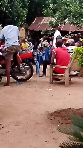 The video shows an outdoor gathering of people. In the foreground on the left, a person wearing a light blue shirt and brown shorts sits on a red motorcycle. To the right, a person in a red shirt sits in a wooden chair. In the background, several people are seated on blue chairs. A building with a corrugated metal roof is visible behind them. The ground is dirt. Green foliage surrounds the area.