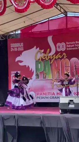 Three young girls are performing a traditional dance on a pink stage in front of a backdrop that reads "Gebyar Panggah". They are wearing colorful costumes with white, purple, and gold accents, and matching headpieces. The girls are moving in synchronized motions, with arm gestures and footwork that are consistent with traditional dance forms. The backdrop indicates the event is related to Indonesia's 80th anniversary, with text reading "Bersatu Bersama Rakyat Sejahtera Indonesia Maju." The stage is set outdoors, under what appears to be a canopy providing shade.