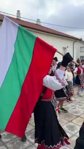 The video shows a crowd of people gathered in an outdoor setting, likely a town square. Many individuals are dressed in traditional Bulgarian folk costumes, including ornate vests and skirts. Some wear elaborate masks with animal-like features and headdresses. A woman in the foreground holds a large Bulgarian flag, waving it as she moves through the crowd. Other flags are visible throughout the group. There are buildings surrounding the square, with light-colored walls and brown roofs. The sky is overcast. The overall atmosphere suggests a festive or celebratory event related to Bulgarian culture and tradition.