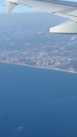 This is a vertical, aerial view of a coastline with a city and a portion of an airplane wing in the foreground. The ocean occupies the bottom half of the frame, appearing a deep, calm blue with a few scattered clouds. A sandy, light-brown coastline runs horizontally across the middle of the image, separating the sea from the land.

Beyond the coastline lies a densely populated urban area. Numerous tall, modern buildings, primarily white or light-colored, are clustered together, forming a cityscape. The buildings vary in height and design, suggesting a significant city. The land behind the city appears to be a mix of developed areas with roads and smaller buildings, interspersed with patches of greenery and some distant hills or mountains.

The top portion of the image features the wing of an airplane, which is white and extends from the left side of the frame towards the right. The wing is angled slightly upwards, showing its structure and a small antenna or sensor protruding from its edge.

The scene is illuminated by sunlight, indicating it is daytime. The sky above the clouds is a pale blue. There are scattered white clouds both near the horizon and as small, wispy formations over the sea. The overall impression is one of flight and observation from above, capturing a coastal city bathed in daylight. There is no visible text in the media file.
