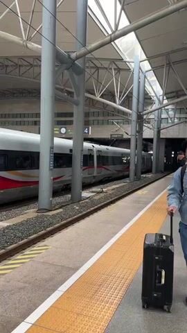 The video shows a train station platform in China. People with luggage are walking around. A high-speed train is stopped at a platform. A man is walking along the platform, pulling a suitcase. An overhead sign indicates that platforms 2 and 3 are straight ahead. An automated message can be heard in the background.