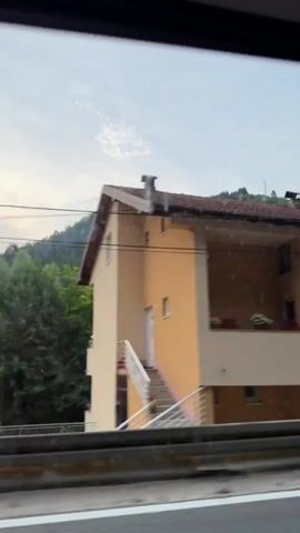 This is a video taken from a vehicle, likely a car or bus, traveling along a road. The view is through a rain-streaked window, indicating it's a wet or overcast day. The scene features a series of houses and buildings lining the roadside, with a backdrop of lush green hills. The architecture appears to be residential, with multiple stories, balconies, and tiled roofs. Some buildings are painted in light colors like beige and yellow, while others are in shades of pink and orange. Several buildings display flags, notably the flag of Bosnia and Herzegovina. The time of day appears to be daytime, possibly late afternoon or early evening, given the muted light. The overall atmosphere is one of passing through a residential area, possibly a town or village, during unfavorable weather. There are no people clearly visible in the video, and the focus is on the passing scenery and the buildings. The activity is the movement of the vehicle, capturing the changing view of the landscape.