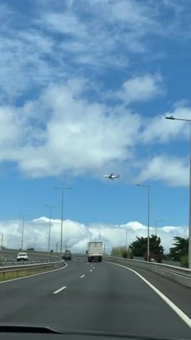 A driver, presumably, records a video while driving on a highway. The highway is two lanes on each side, with a guardrail separating the sides of the highway. There is a white truck on the highway in front of the driver, and another car. The highway is curving. There are street lights along the highway. In the distance, over the highway, a passenger airplane is descending, presumably to land. The sky is light blue with white clouds.