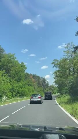 A vehicle is driving on a paved road flanked by lush green trees and vegetation on both sides under a clear blue sky with some scattered white clouds. Two cars are visible ahead, a silver hatchback followed by a black minivan. The camera is positioned from inside the vehicle, possibly from the driver's perspective, capturing the view through the windshield.