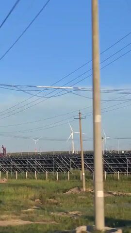 The video shows a landscape with a clear blue sky. In the foreground, there are grassy fields and several utility poles with electrical wires running between them. In the middle ground, there is a large array of solar panels, tilted and arranged in rows. Behind the solar panels, there are several wind turbines with their blades visible. There are also some structures that appear to be industrial, possibly oil pumps, visible in the distance. The camera appears to be moving, giving a sense of travelling through the scene. There are no people clearly visible in the frame. The environment suggests a location that is harnessing renewable energy.