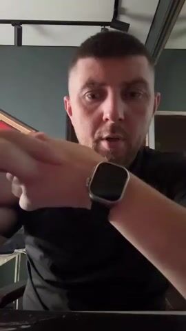 The video features a close-up shot of a man's face and wrist, displaying a smartwatch. The man is wearing a black polo shirt and a silver ring on his left hand. His hair is short and styled, and he has a light beard. He is looking directly at the camera with a neutral expression.

The smartwatch on his wrist has a square screen and a dark gray band. In the first few seconds, the watch displays a bright orange screen with circular icons, suggestive of a weather or a notification interface. The man is seen touching the watch face, and the screen changes to a different display showing a circular analog clock face with a red second hand and a digital time display, along with what appear to be notification or app icons.

The background is out of focus but suggests an indoor setting. The wall behind the man is a dark teal color, and there's a portion of what looks like a framed picture or decorative item with red and gold elements visible on the left side of the frame. The lighting appears to be from an artificial source, likely indoors, and the time of day cannot be determined.

The activity taking place is the man demonstrating or interacting with his smartwatch, likely showing its features or a specific application. His focused gaze and hand gestures indicate he is actively engaged with the device. No external weather cues are visible. The man's expression is calm and direct, without any strong emotional display. There is no visible text on the man's clothing or the background elements. The location context provided (Protasovo, Russia) is not visually evident from the image itself.