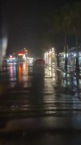 The video shows a wet street at night, likely after rain, with streetlights and decorative lights reflecting on the wet asphalt. Cars are driving by, their headlights and taillights creating streaks of color on the road. On the right side of the street, palm trees are decorated with string lights, and there are some signs displayed. The overall atmosphere is dark and moody, with the lights creating a sense of vibrancy against the night sky.
