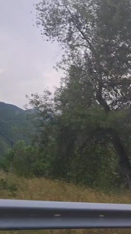 The video displays a scenic view of lush green mountains with a cloudy sky overhead. The perspective is from a moving vehicle, indicated by a metal guardrail in the foreground and the blurred vegetation along the roadside. The camera pans from left to right, revealing layers of mountains receding into the distance, with a valley visible between them. The vegetation on the mountains is dense and appears to be a mix of trees and shrubs. The overall atmosphere is serene and natural.
