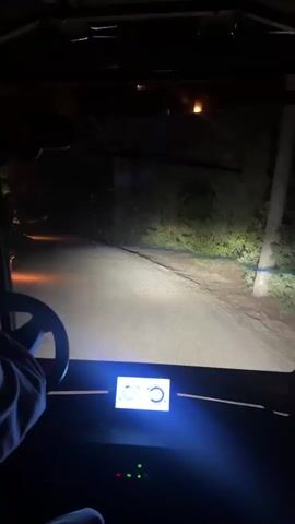 This media file captures a first-person perspective of driving a golf cart at night on a winding, unpaved road. The scene is illuminated by the golf cart's headlights, which cast a bright beam ahead, and by scattered outdoor lighting that appears to be a mix of yellow and orange hues, suggesting lamps or torches along the path. Dense vegetation flanks the road, creating a dark and somewhat mysterious atmosphere.

The primary subject is the act of driving, with a hand visible on the steering wheel of the golf cart. In the foreground, a digital display panel on the golf cart glows with a blue light, showing abstract shapes and what could be indicators or a speedometer. The overall setting appears to be a rural or natural area, possibly a resort or a private estate, given the golf cart and the decorative lighting. The time of day is clearly nighttime, and the weather is not discernible from the visual information. There is no visible text within the media file. The mood is calm and perhaps slightly adventurous, given the nocturnal drive through a dimly lit, natural environment. The location context provided indicates Grândola, Portugal, which aligns with the possibility of a rural or semi-rural setting.
