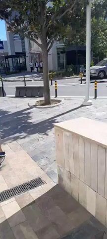 A video shows a person's legs and feet from the knees down. The person is wearing blue and white athletic shoes and is sitting on a metal chair. The ground is paved with large, rectangular stone tiles. A metal grate is visible in the pavement, likely a drain. Shadows from trees are cast across the ground, indicating it is daytime and sunny. The scene appears to be an outdoor public space, possibly a street or plaza.