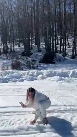 The video shows a woman in a white jumpsuit squatting in the snow, forming snowballs. She then stands up and throws the snowballs into the air while holding a phone in her hand. The background is a snowy landscape with a forest of tall, bare trees. It is a sunny day.