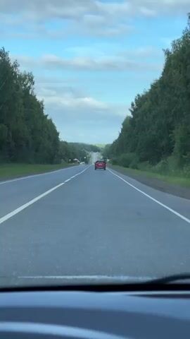 A first-person perspective video shows a view from inside a car driving down a two-lane asphalt road. The road is bordered on both sides by dense green trees. In the distance, the road curves and dips down a hill. A red car is visible ahead, also driving down the road. Several traffic signs are present along the roadside. The sky is blue with scattered clouds.