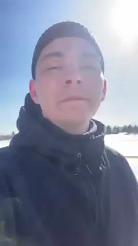 A person wearing a black beanie and black jacket stands in front of a landscape with snow and trees. The sun is shining brightly in a clear blue sky. The person looks at the camera.