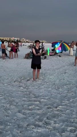 A young person is juggling glowing objects on a sandy beach at dusk. The person is wearing a dark t-shirt and shorts. Several people are visible in the background, sitting on chairs, standing, and walking. A colorful umbrella is set up on the beach. The sky is cloudy and overcast. The glowing objects appear to be square or rectangular and are illuminated with green, blue, and pink lights.