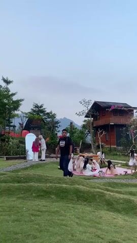 The media file captures an outdoor gathering at a scenic location, likely a park or resort, in Kesiman, Indonesia. The scene is set on a gently sloping grassy hill with a modern wooden building in the background. A group of people, including several women in hijabs and a man in a black t-shirt, are engaged in a picnic. They are seated on pink and white checkered blankets spread on the grass. In the foreground, a young girl with dark hair and a white shirt is eating something and looking towards the camera, her expression suggesting a moment of casual enjoyment. Further back, another young girl in a white dress stands near a large, abstract white structure with an oval opening.

The time of day appears to be late afternoon or early evening, with soft, diffused light suggesting overcast conditions. The sky is a pale grey, and distant mountains are visible in the background, adding to the natural beauty of the setting. The overall atmosphere is relaxed and convivial, with people enjoying leisure time together outdoors.

Visible text includes "FLOR VERDE" written on a light-colored building, possibly indicating the name of the establishment or venue. The presence of picnic blankets, food (implied by the girl eating), and the gathering of people signifies a leisurely picnic or a similar social event. The modern architecture of the building contrasts with the natural landscape, creating an interesting visual juxtaposition. The image conveys a sense of peace and a pleasant day spent in a visually appealing environment.