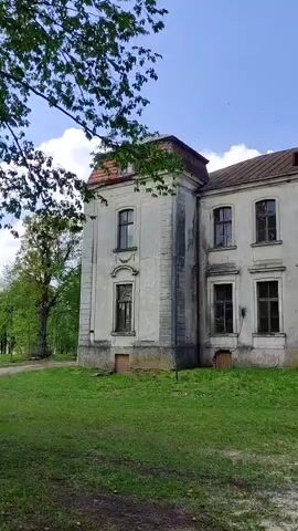 The video shows a slow panning shot of a large, old, and dilapidated building with a baroque style. It has multiple stories and many windows, some of which are broken or boarded up. The building is painted a light color, but the paint is peeling and there are signs of disrepair. There is a grassy lawn in front of the building and some trees nearby. The sky is blue with some white clouds. A child's voice can be heard in the background, speaking in a language other than English.