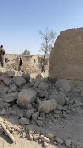The video shows a landscape of destroyed buildings and rubble, under a clear blue sky. The area appears arid. Several men dressed in traditional clothing are present, standing amongst the debris. Some are observing the scene, while others are walking through the wreckage. The buildings appear to be made of a light-colored material, possibly mud brick, and are partially collapsed. The ground is covered in rocks and debris.