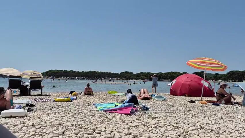 A wide shot of a crowded beach on a clear, sunny day. The beach is covered in white pebbles, and people are spread out across it. Some are lying on towels or blankets, others are sitting in chairs or under umbrellas. Several tents are set up on the beach, providing shade. The ocean is visible in the background, with many people swimming and enjoying the water. A line of trees runs along the distant shore. The overall impression is a lively and active beach scene.